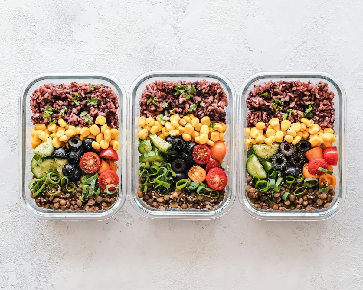 Drei Meal-Prep-Boxen mit Reis, Mais, Oliven, Tomaten und Linsen